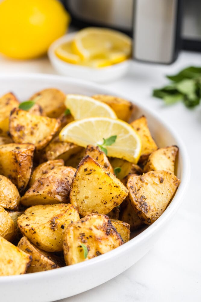 side view of greek potatoes in a white bowl, garnished with two slices of lemon and fresh oregano. there's an air fryer in the background