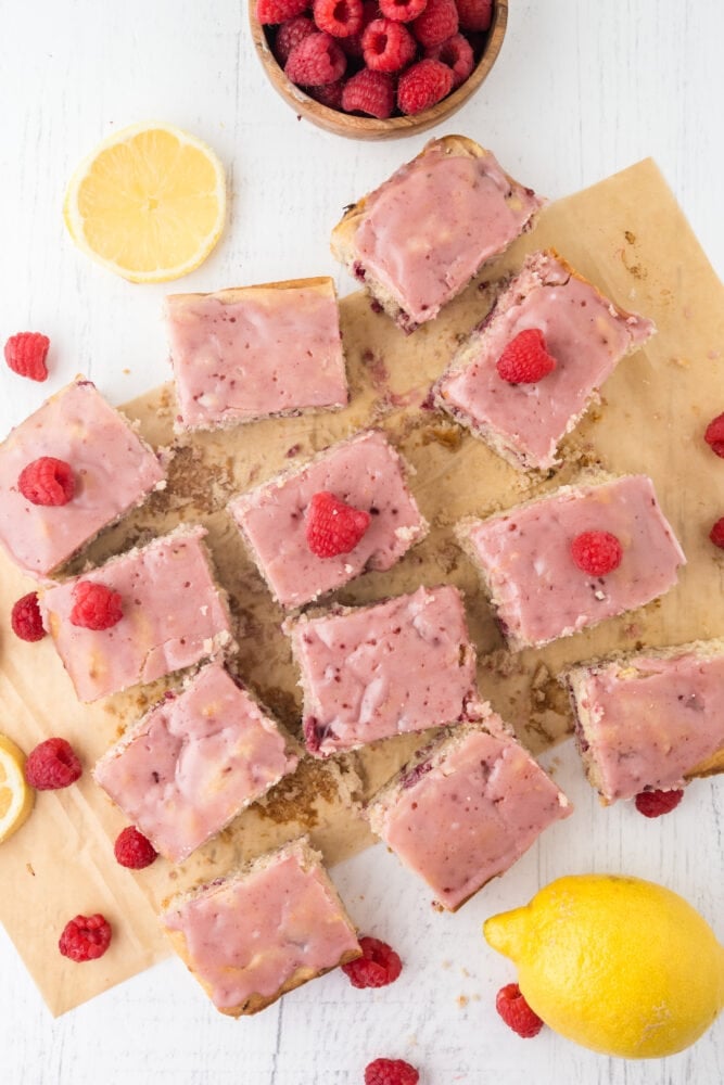 Raspberry lemon bars cut into 12 squares and garnished with pink raspberry glaze, fresh raspberries, and lemon slices off to the side.