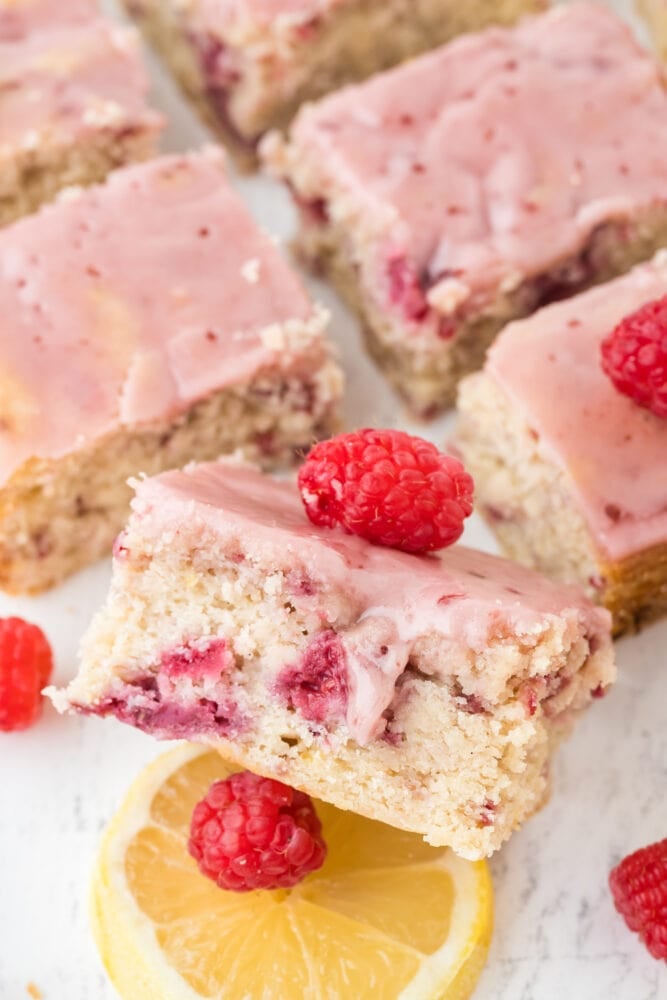 raspberry lemon bars cut into squares, with one tilted up on its side being held up by a raspberry and lemon slice, to show the delicious cakey texture