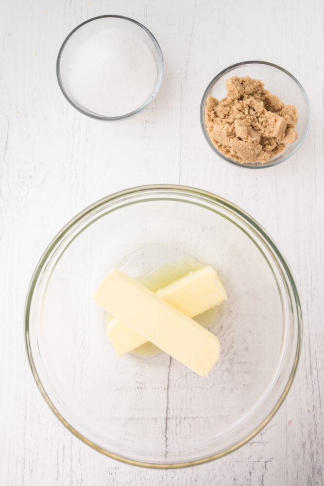 two sticks of butter, softened, in a glass bowl. There's a mini glass bowl of sugar, and also one with brown sugar, ready to mix with the butter