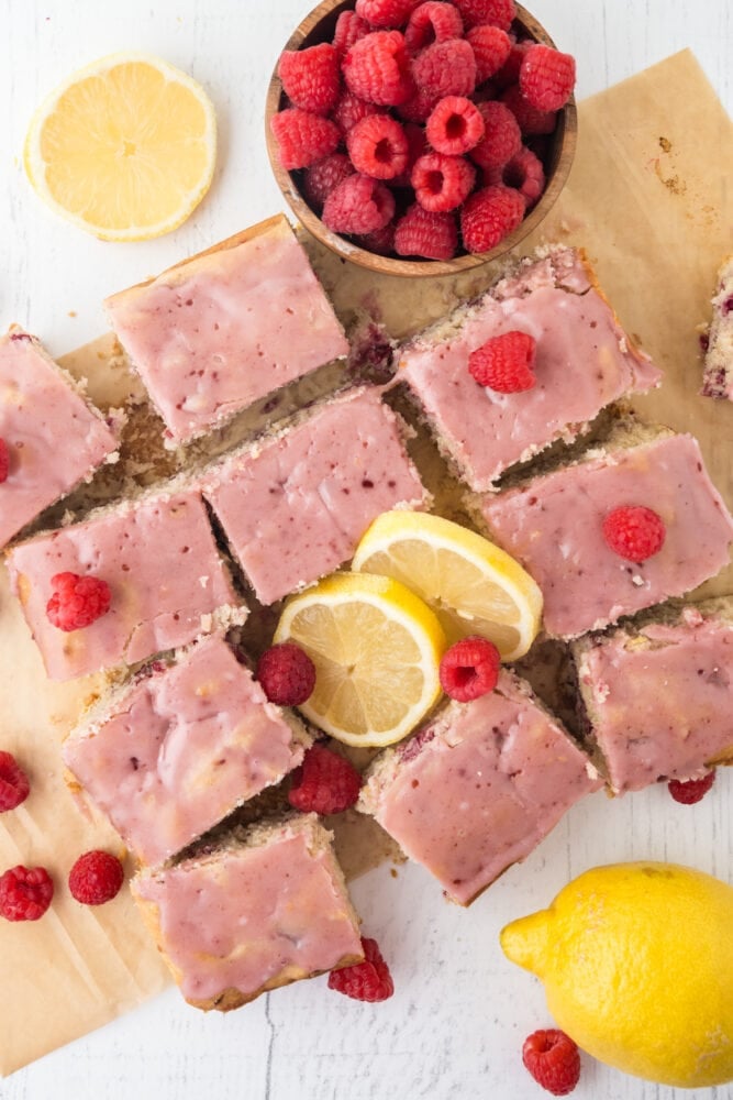 Raspberry lemon bars cut into 12 squares and garnished with pink raspberry glaze, fresh raspberries, and lemon slices off to the side. There's also a wood bowl of fresh raspberries