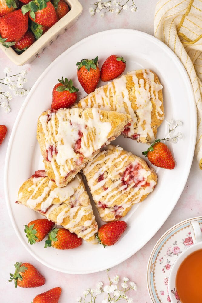strawberry scones on a white oval plate, drizzled in white sugar glaze, and garnished with fresh strawberries and white flowers.