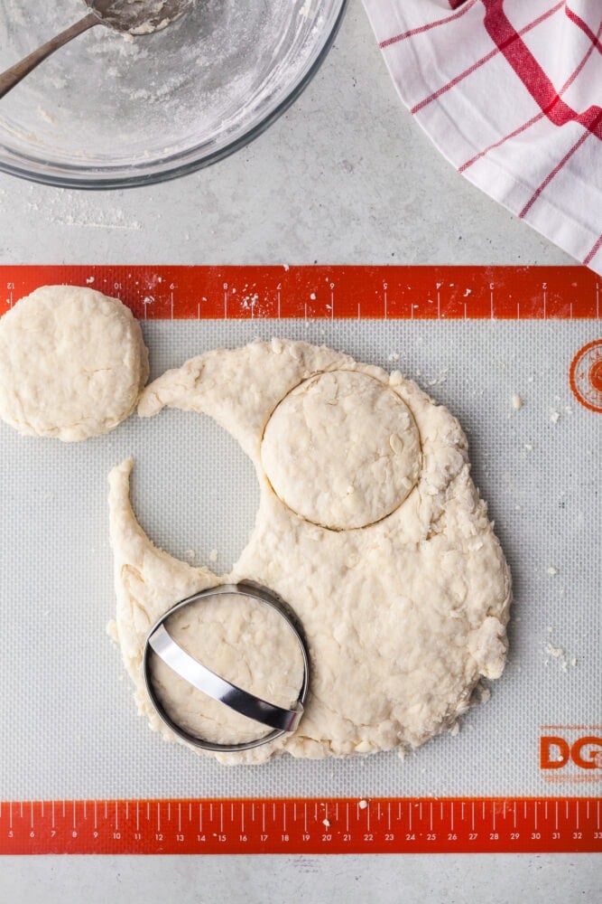 Biscuit dough rolled out onto a silicone sheet, there's one biscuit cut out with a biscuit cutter, and another in a round shape that was just cut.