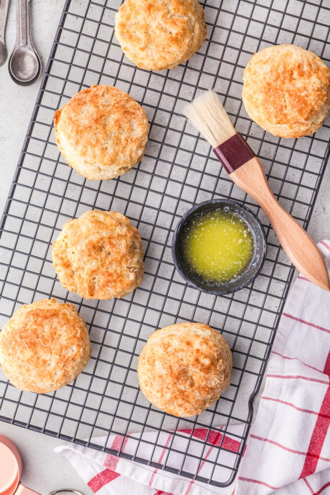 Six air fryer biscuits cooling on a wire rack, with a small bowl of melted butter and pastry brush to the side.