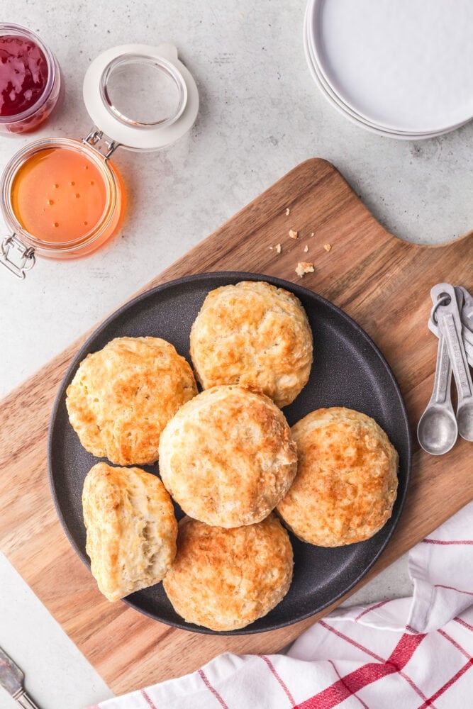 A round black plate holding six golden biscuits. The plate is on a wood cutting board and there's a peach and strawberry jam to the side ready for serving. There's also a white and red checkered towel and measuring spoons.