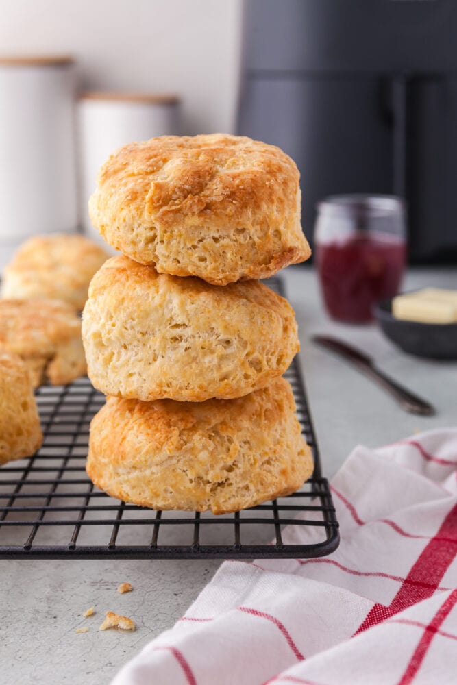 A stack of three biscuits on a wire rack. Behind the biscuits are an air fryer, white flour and sugar canisters, a black air fryer, jar of berry jam, and little serving dish of butter.