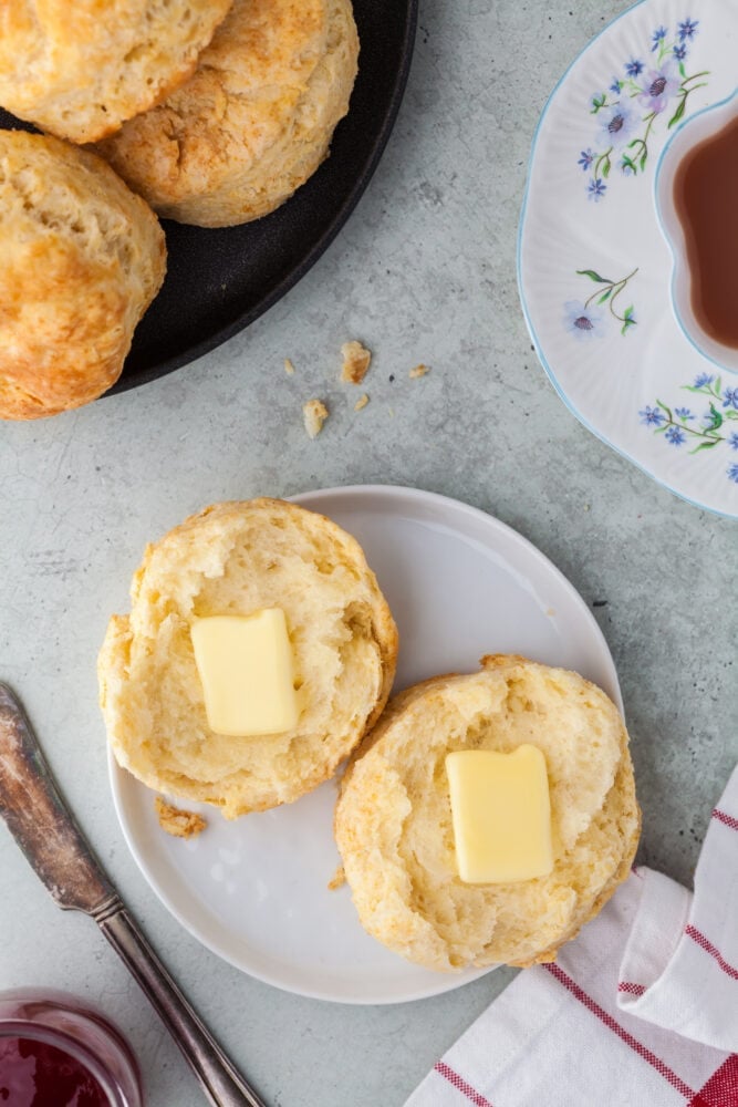 A biscuit cut in half on a white plate with a square of butter on each side. There's a cute white tea cup with blue flowers to the side as well as an extra plate of biscuits.
