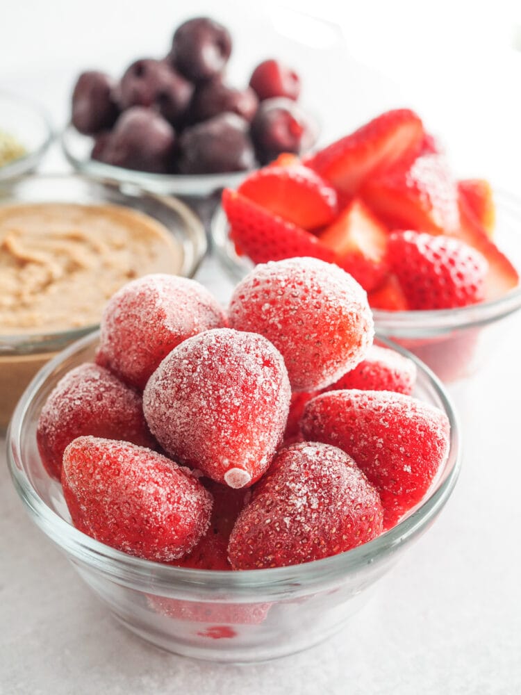 Frozen strawberries in a glass bowl with other protein shake ingredients in the background.