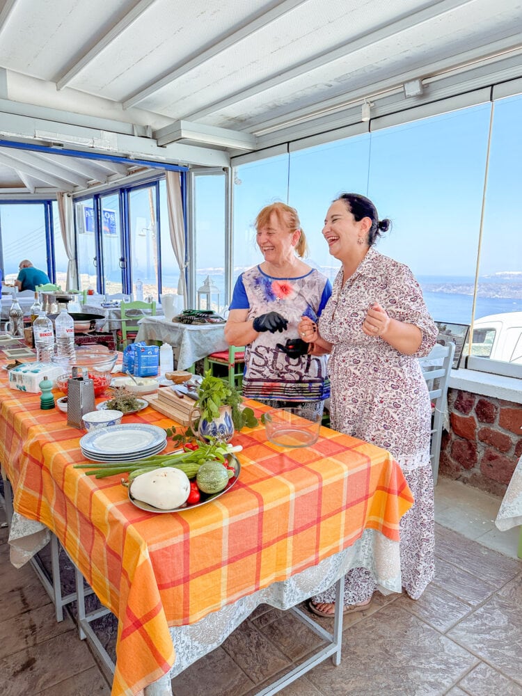 Two women laughing as they demonstrate Greek cooking techniques in Santorini.
