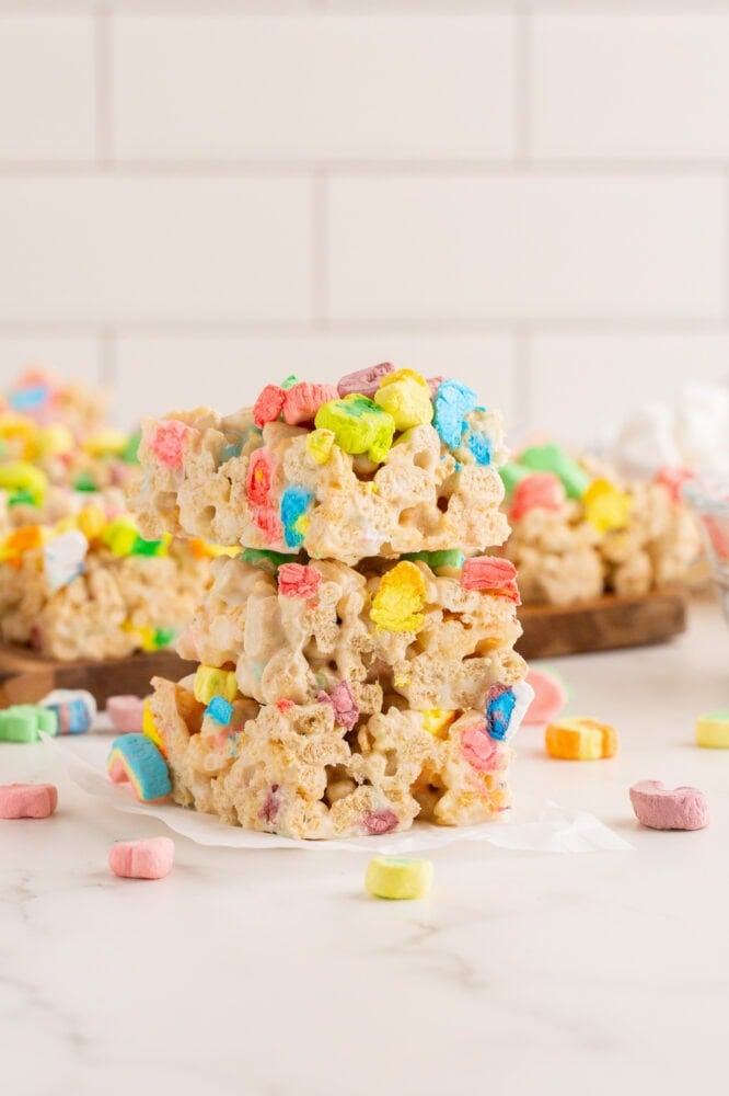 Side view of a stack of of lucky charms rice krispie treats sitting next to a cutting board full of treats.