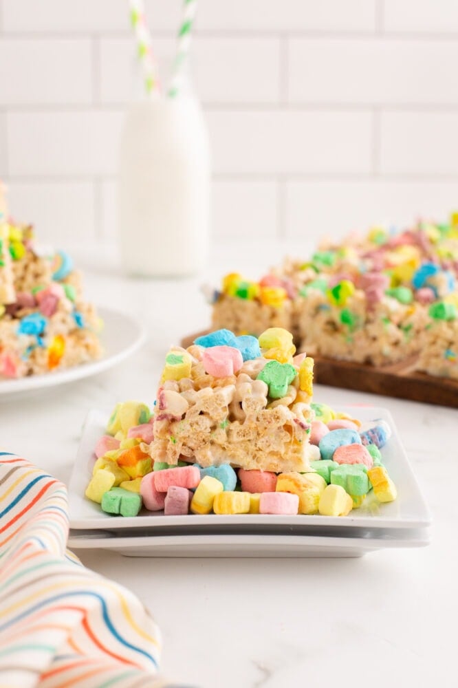 Lucky charms rice krispie treat sitting on extra lucky charms marshmallows on a white plate. Theres a jar of milk with green and white striped straws, and a cutting board with more treats in the background.