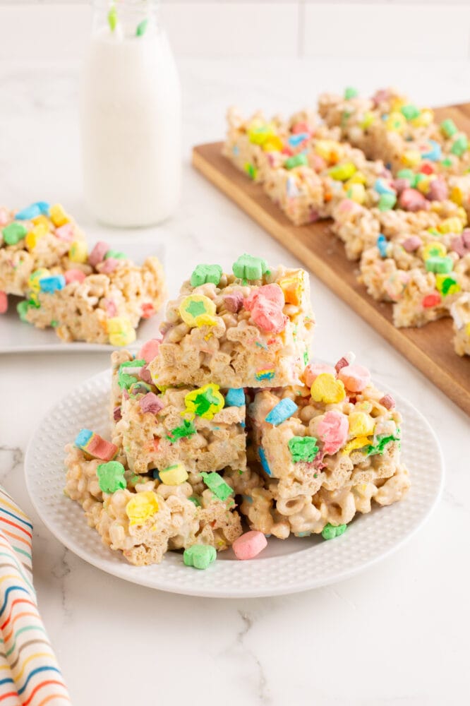 Side view of a white plate with a stack of of lucky charms rice krispie treats sitting next to a cutting board full of treats and a glass jar of milk. There's also a colorul rainbow striped napkin to the side that matches the lucky charms.