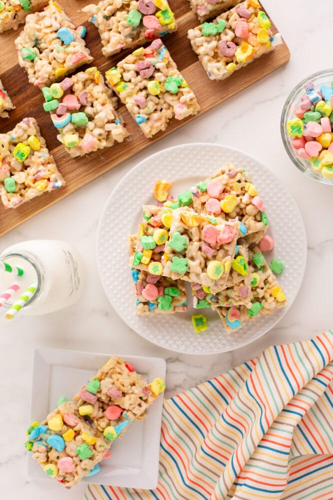 Overhead look at a white plate with a stack of of lucky charms rice krispie treats sitting next to a cutting board full of treats and a glass jar of milk. There's also a colorul rainbow striped napkin to the side that matches the lucky charms.