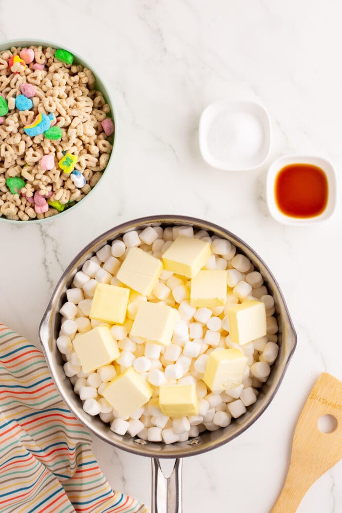 Stainless steel saucepan with mini marshmallows and 10 dabs of butter on top, before being melted. There's a bowl of Lucky Charms cereal and a wood spoon off to the side.