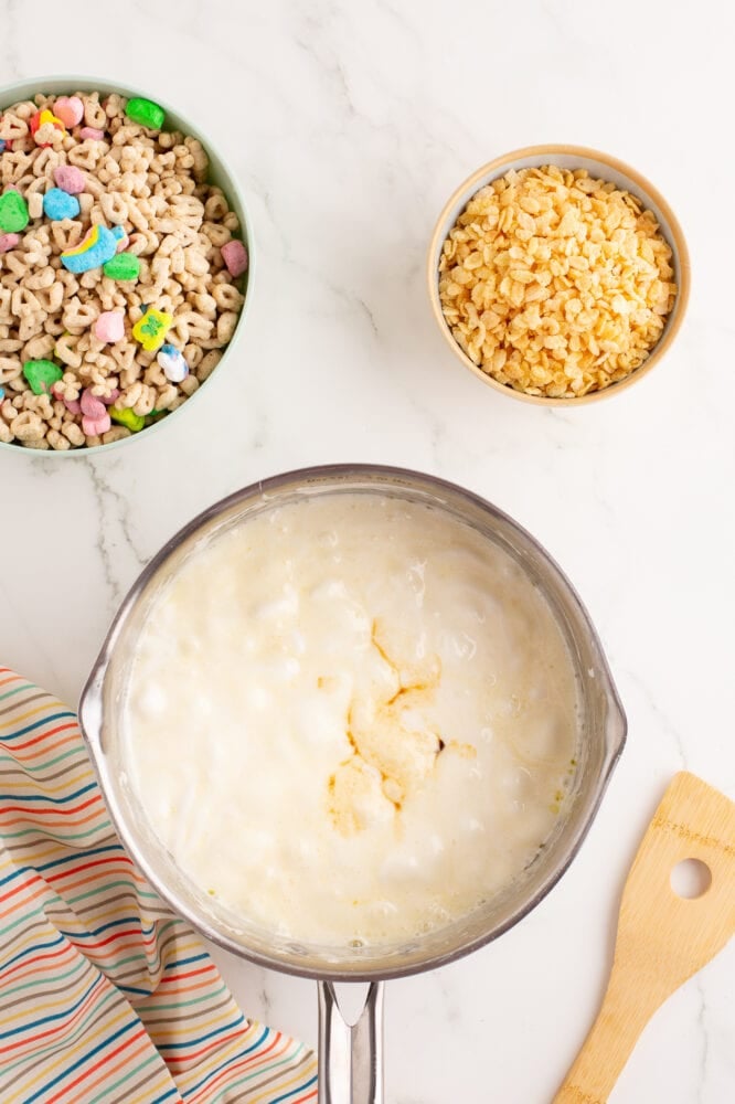 Melted butter and marshmallows in a stainless steel pan, with vanilla and salt added on top. There's a bowl lucky charms cereal and rice krispie cereal set to the side ready to mix in with a wood spoon.