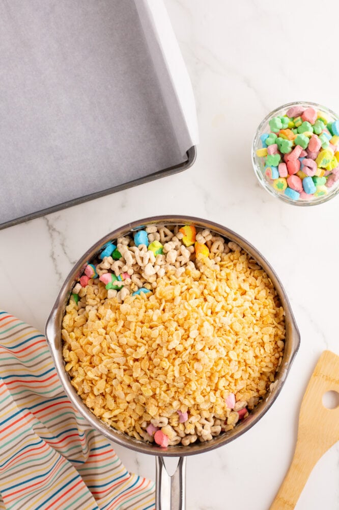 Lucky charms rice krispies mix in a steel pot, with parchment lined 9x13 bake pan to the side and a small bowl of extra charms marshmallows to add on top. The parchment paper goes over the sides of the pan to make it easy to lift the treats once they are cooled.