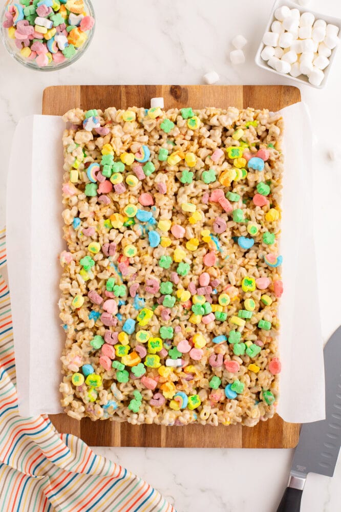 Chilled lucy charms rice krispie treats on parchment paper resting on a cutting board before getting cut to serve. There's a knive to the side as well as extra bowls of marshmallows.