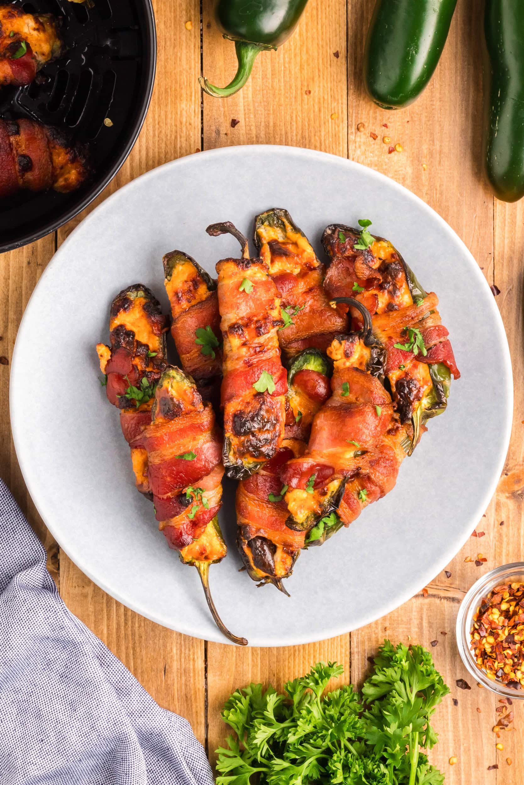 A white plate on a wood picnic table. The plate has bacon wrapped jalapeno poppers stacked and sprinkled with parsley. They look delicious.