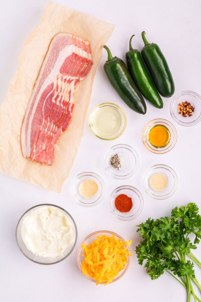 Ingredients for air fried bacon wrapped jalapeno poppers including bacon, jalapenos, honey, salt, pepper, garlic powder, paprika, onion powder, cream cheese, shredded cheddar cheese, avocado oil, and parsley.