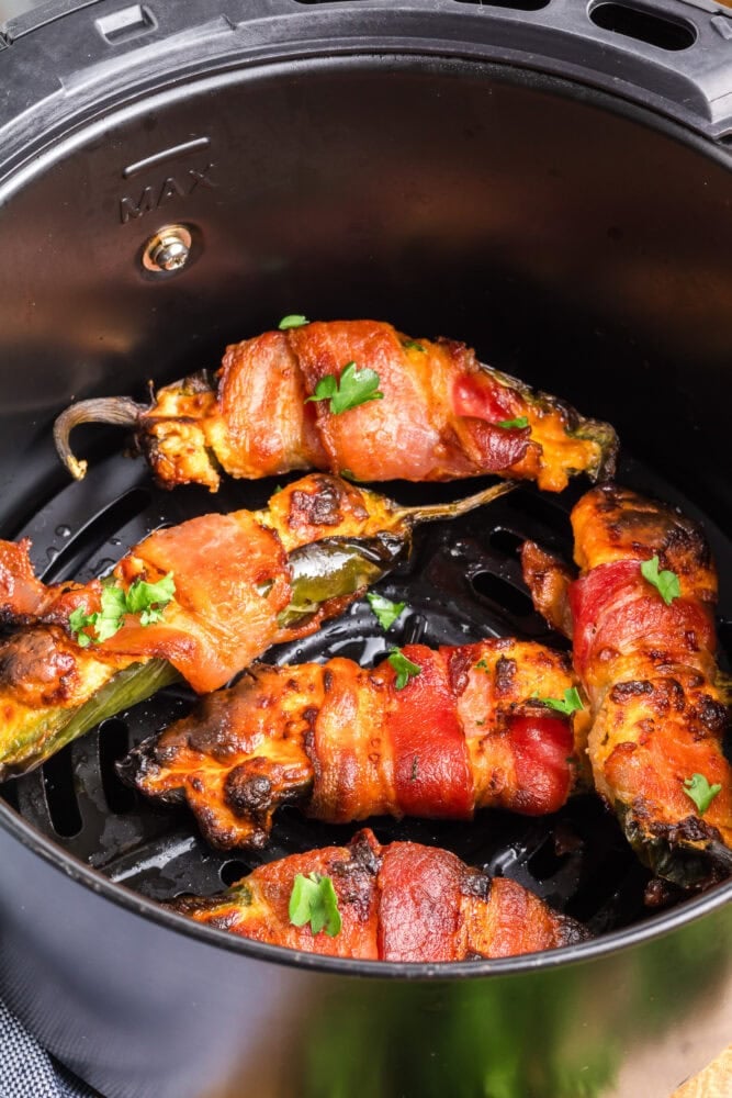 Air fried bacon wrapped jalapeno poppers in an air fryer basket. They look browned and crispy and delicious.