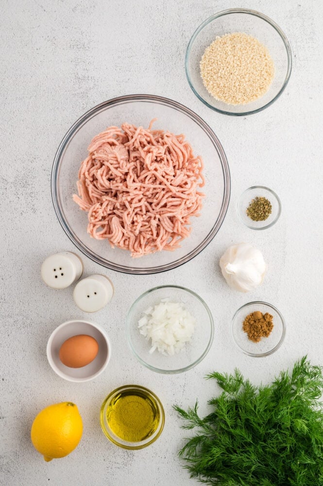 Ingredients for making Greek chicken meatballs including ground chicken, bread crumbs, salt and pepper, garlic, diced onion, cumin, olive oil, an egg, fresh dill, and lemon.