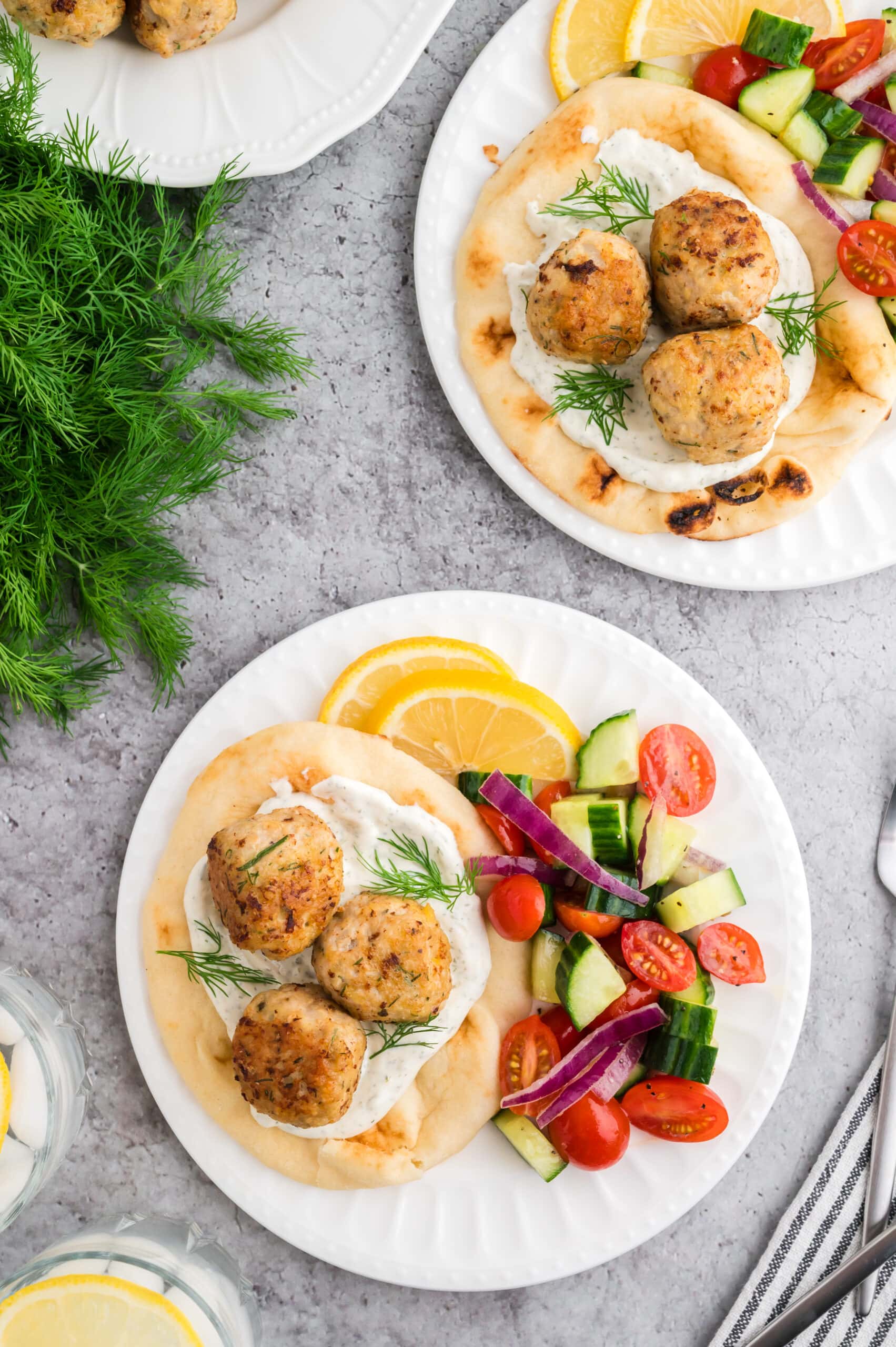 Two white plates on a grey concrete background. The plates each have a pita with tzatziki sauce, three Greek chicken meatballs, and a side Greek salad garnished with two lemon slices. The the side of the dishes is a bunch of fresh dill, a plate with extra meatballs, glasses of water with lemon, and forks for serving.