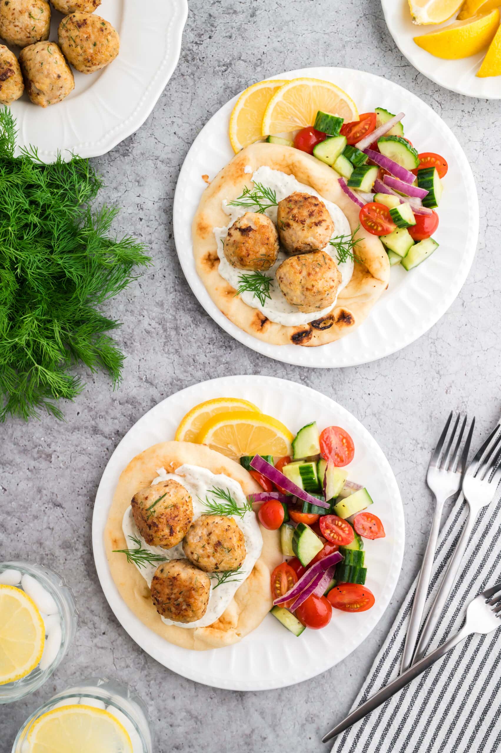 Two white plates on a grey concrete background. The plates each have a pita with tzatziki sauce, three Greek chicken meatballs, and a side Greek salad garnished with two lemon slices. The the side of the dishes is a bunch of fresh dill, a plate with extra meatballs, glasses of water with lemon, and forks for serving.