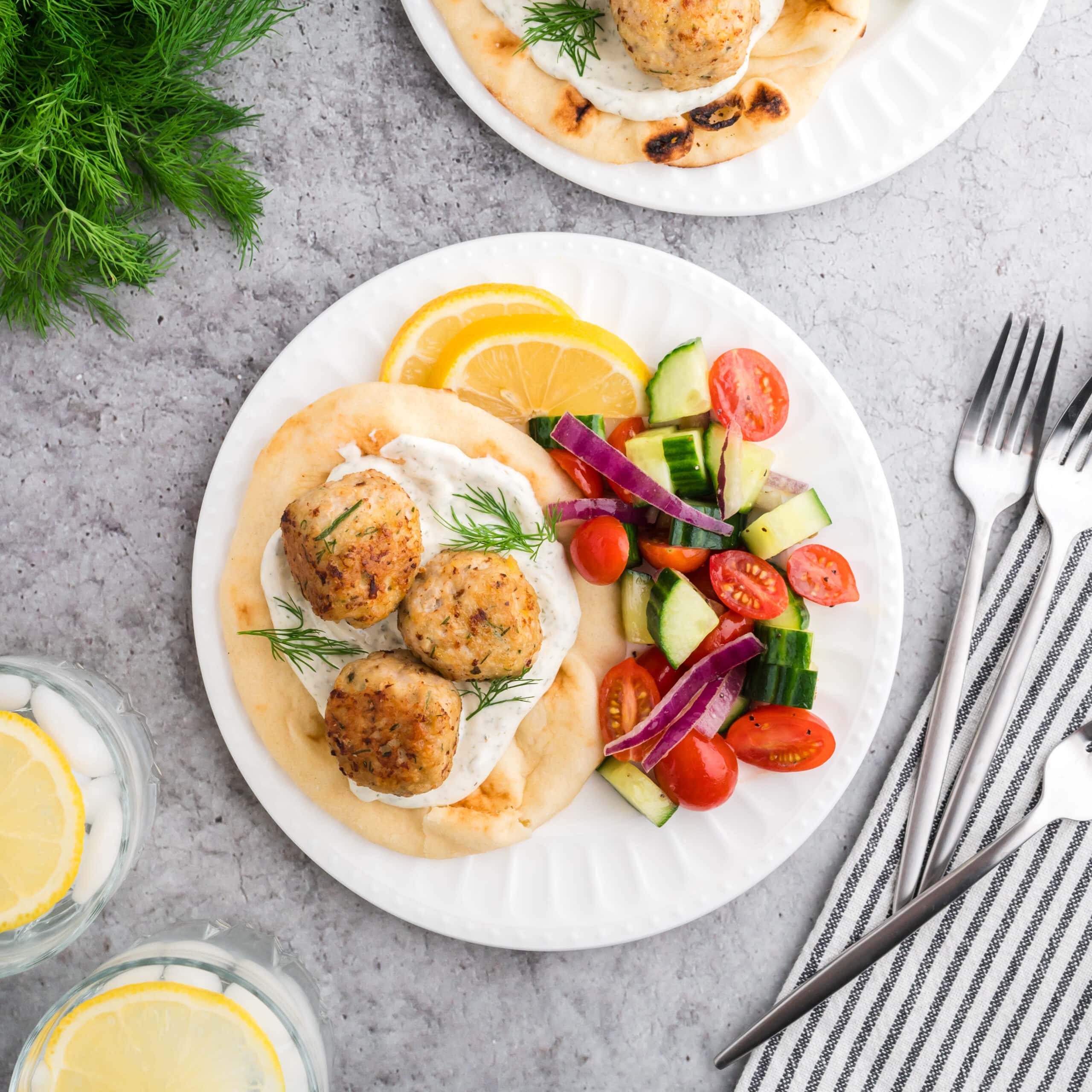 Two white plates on a grey concrete background. The plates each have a pita with tzatziki sauce, three Greek chicken meatballs, and a side Greek salad garnished with two lemon slices. The the side of the dishes is a bunch of fresh dill, a plate with extra meatballs, glasses of water with lemon, and forks for serving.