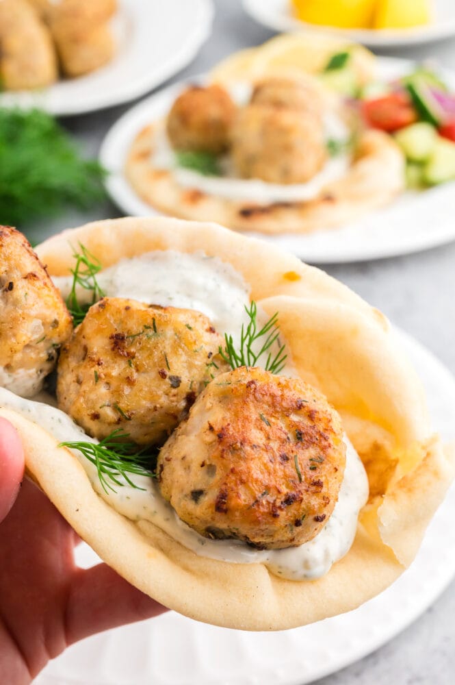 Close up look at three Greek meatballs, golden brown and looking delicious placed on a pita with tzatziki and dill and held in a hand like a taco.
