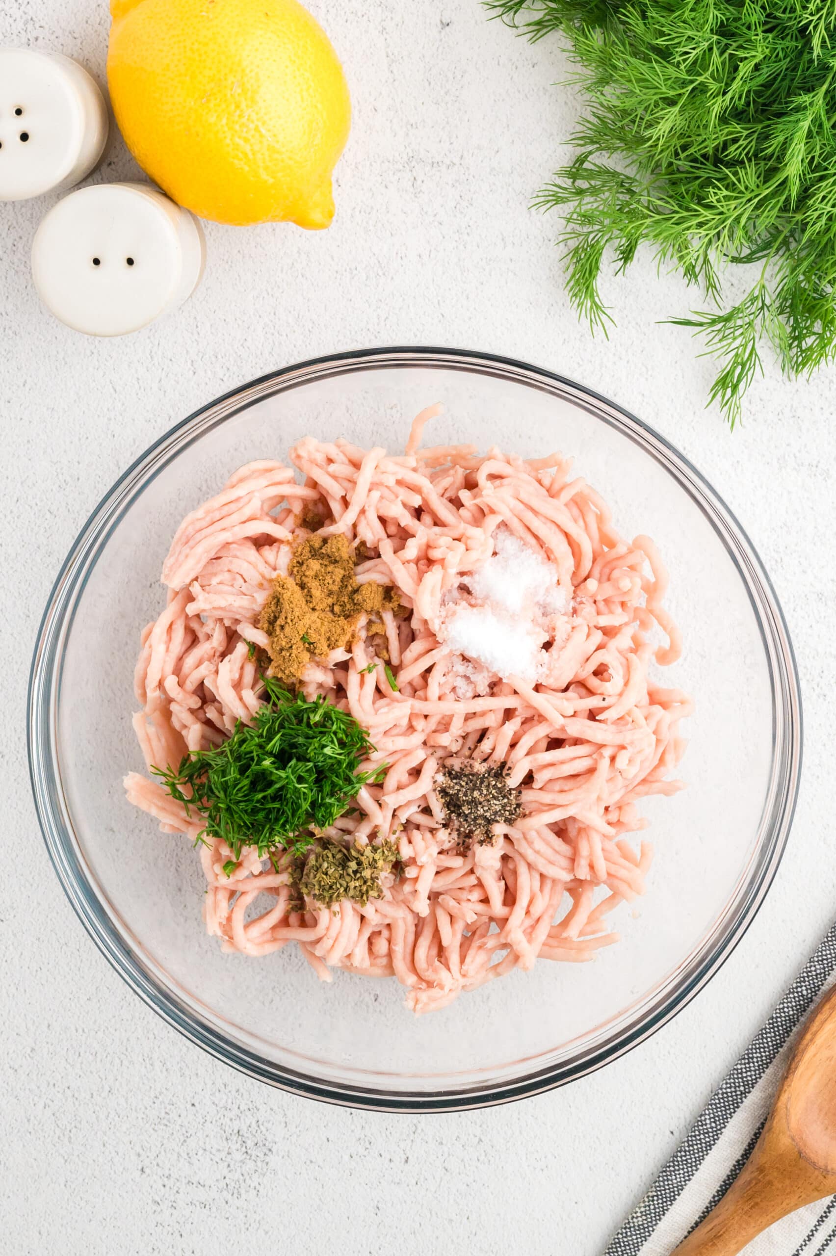 Ground chicken in a clear glass bowl with fresh dill and spices on top before mixing.