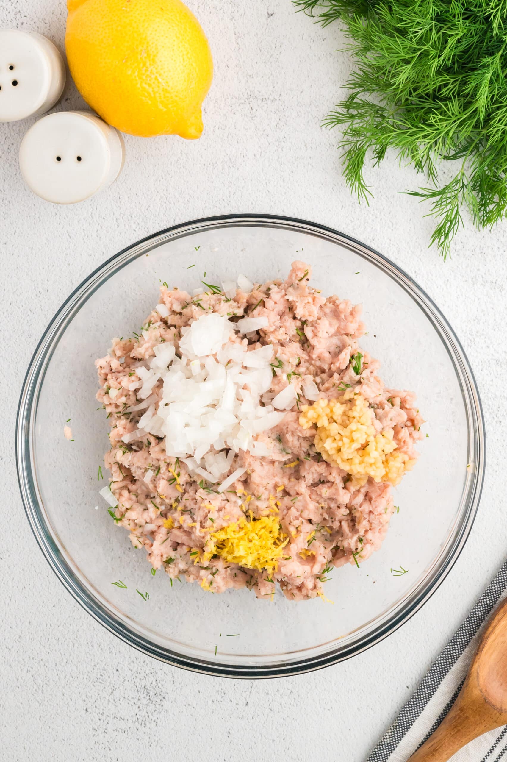 Ground chicken in the process of being mixed in to meatballs, with diced onion, diced garlic, adn lemon zest on top.