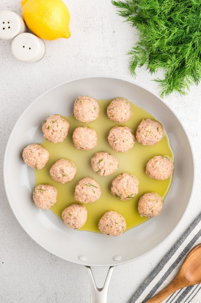 Raw, round Greek chicken meatballs in a frying pan with some oil.