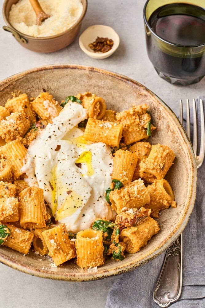 Earthenware bowl with rigatoni pasta, pesto, spinach, and burrata sprinkled with red pepper flakes and olive oil. There's a glass of red wine to the side as well as a bowl of parmesan and a little bowl of red pepper flakes. It looks delicious!