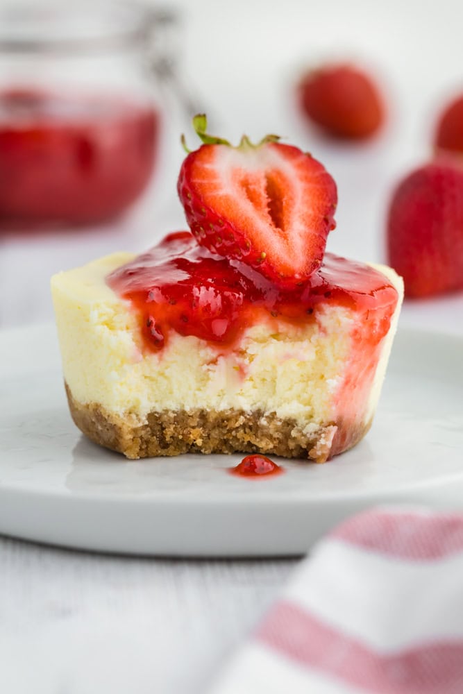 A single mini cheesecake topped with a sliced strawberry, strawberry sauce, and a single bite take out of it.