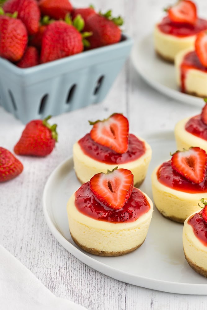 plate of mini cheesecake bites topped with sauce and a sliced strawberry. There's a pint of fresh strawberries in the background.