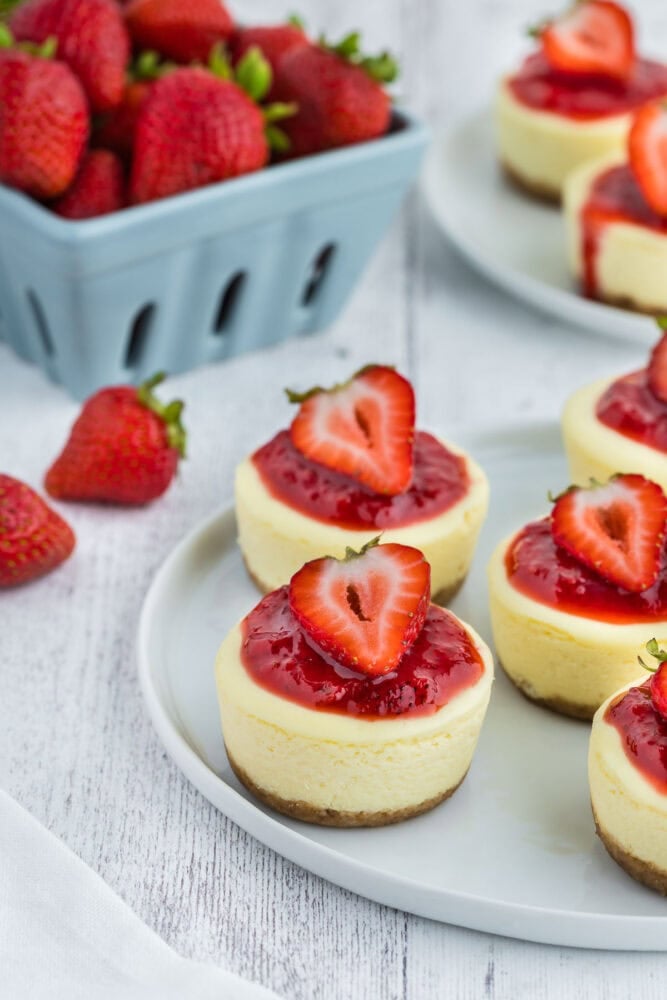 A plate of mini cheesecakes topped with sauce and sliced ​​strawberries.  In the background is a glass of fresh strawberries.