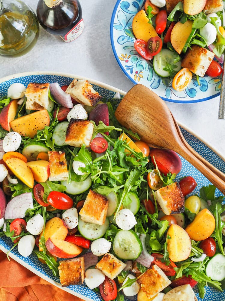 Large serving dish of peach panzanella salad with a small plate to the side as well as small bottles of vinegar and oil.