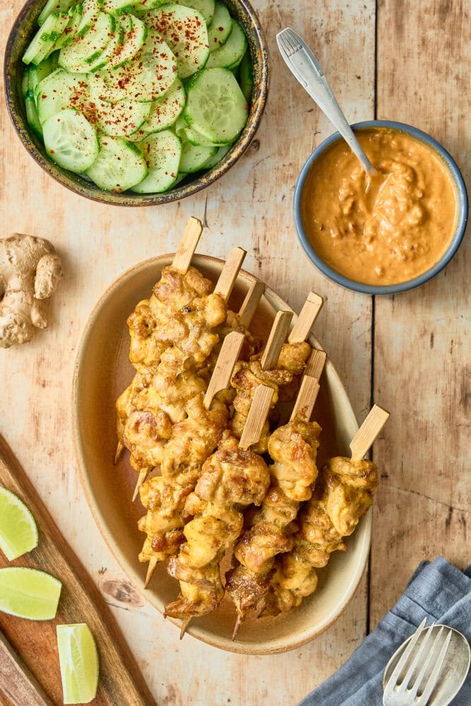 a wood table with a plate of Thai chicken satay on bamboo skewers in an oval tan plate. There's also a bowl of cucumber salad, and a small bowl of Thai peanut sauce to the side.