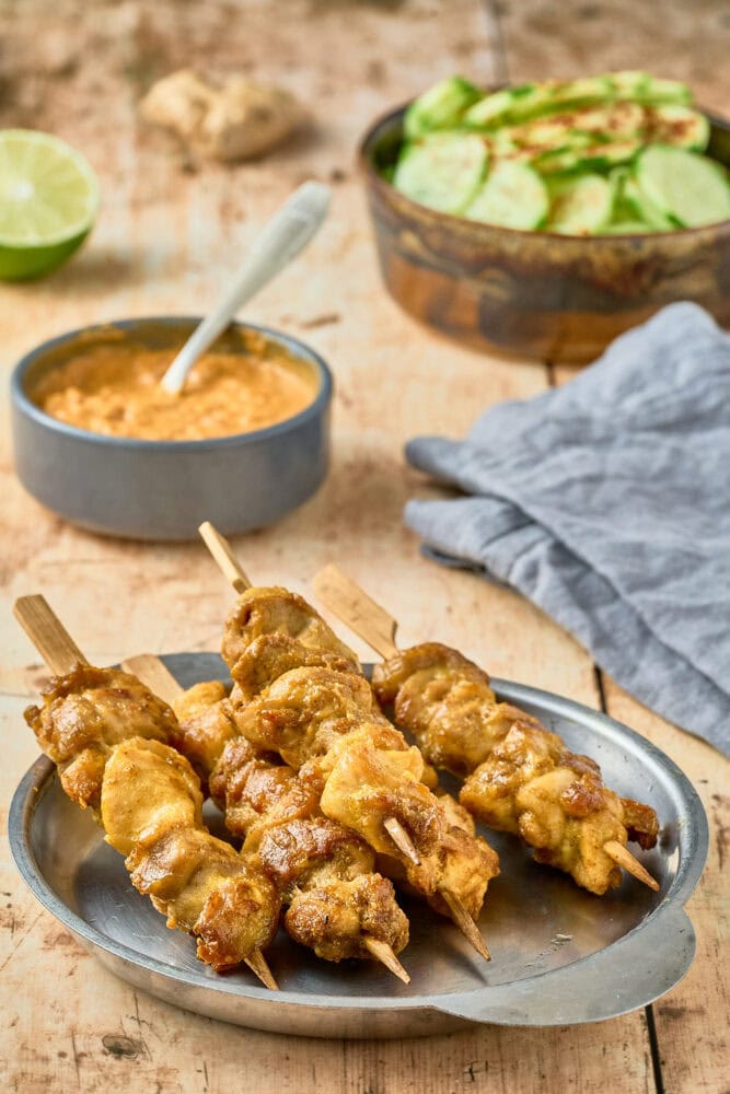 Thai chicken satay skewers, caramelized and cooked, on a metal plate with a napkin in the background along with a bowl of peanut sauce and cucumber salad.