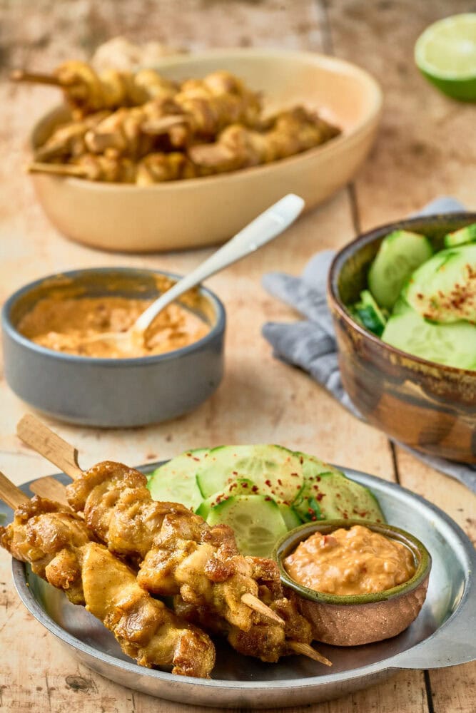 chicken satay plated in a metal bowl with cucumber salad and peanut sauce