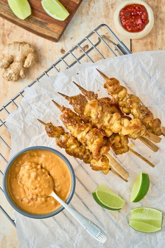 thai chicken skewers and peanut sauce in a bowl resting on a piece of parchment paper with limes