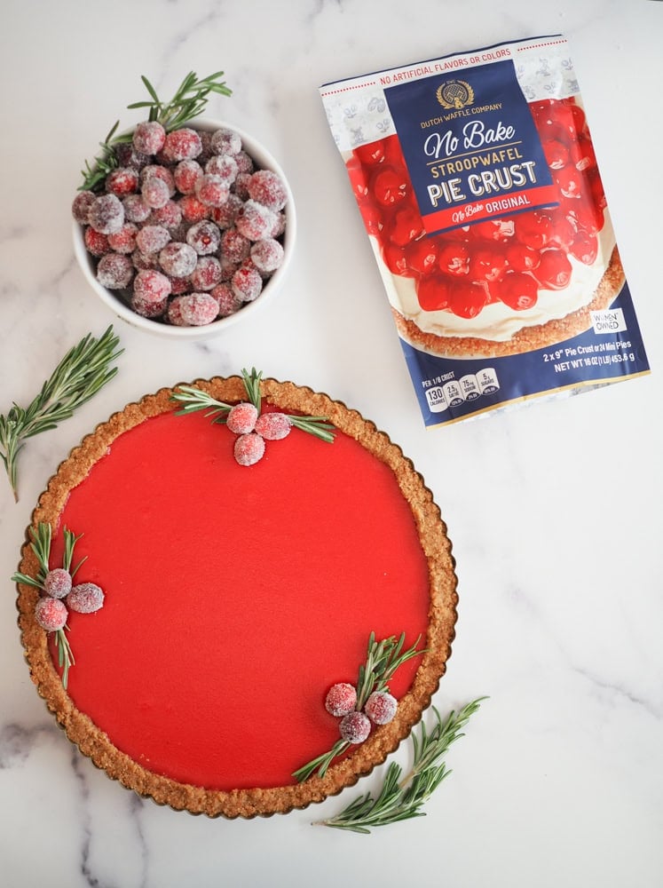 Cranberry tart garnished with sugared cranberries, with a bowl of extra sugared cranberries to the side and a package of stroopwaffel pie crust mix.