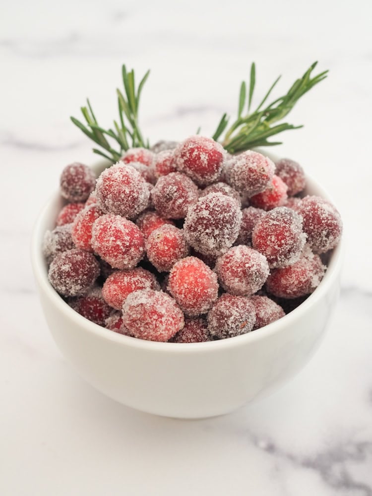 Bowl of sugared cranberries garnished with rosemary. They look like they're covered in glittery snow.