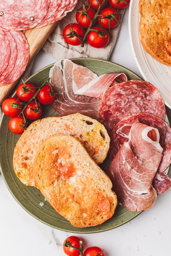 A green plate of pa amb tomaquet, or Spanish tomato bread, with mini tomatoes on the vine, and various charcuterie meats.