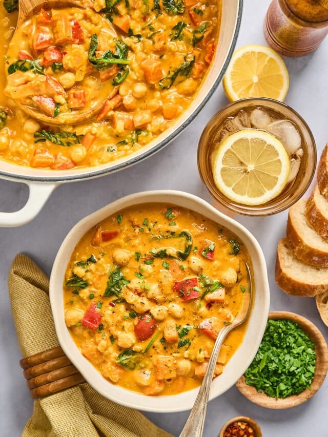 overhead look at a bowl of creamy chickpea stew next to a large white enameled cast iron pot full of it to share, and small dishes of misc garnishes including herbs and lemon.