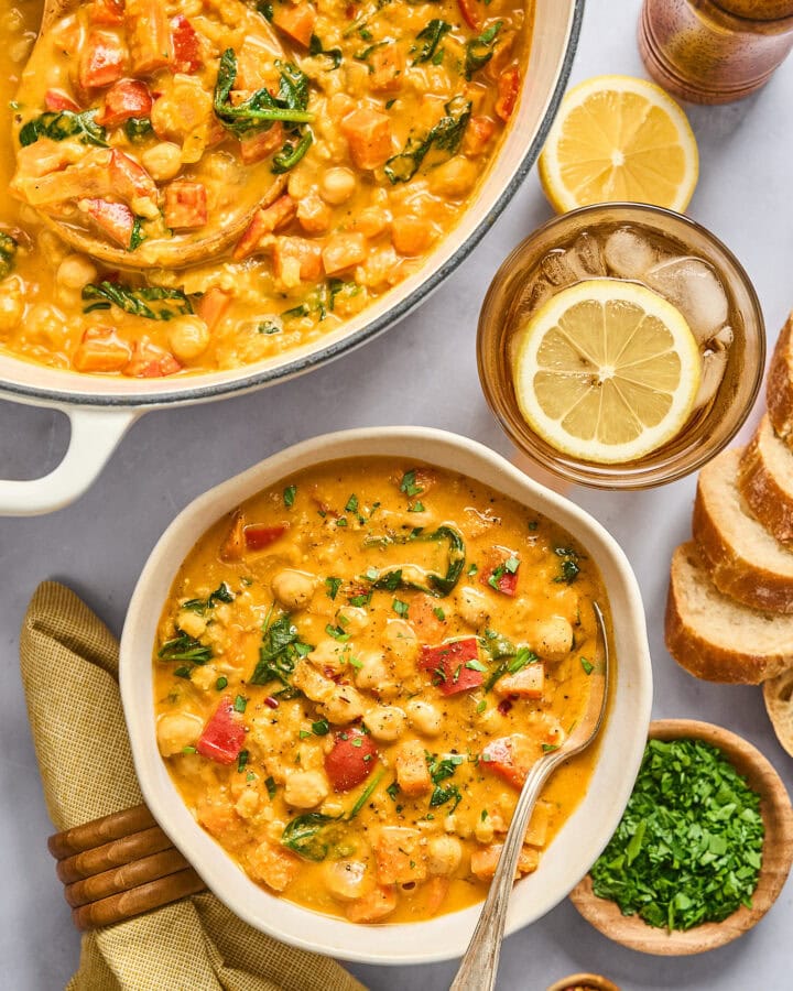 overhead look at a bowl of creamy chickpea stew next to a large white enameled cast iron pot full of it to share, and small dishes of misc garnishes including herbs and lemon.