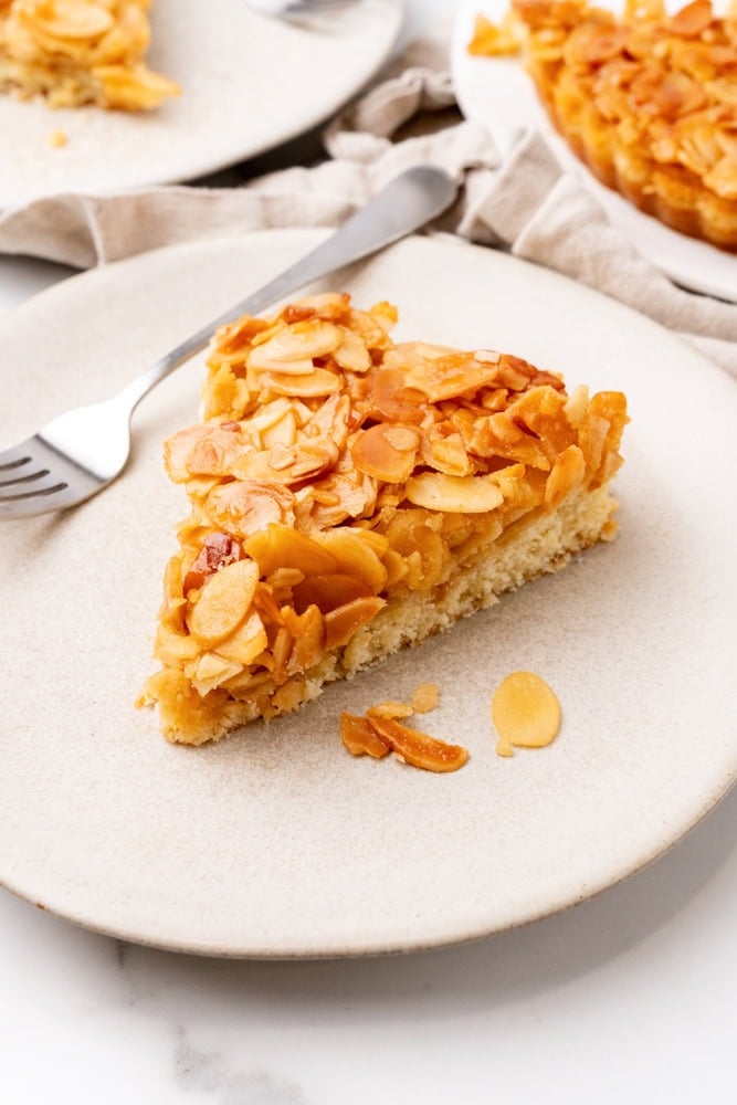 A pie wedge shaped slice of almond tart on a beige plate with a fork.