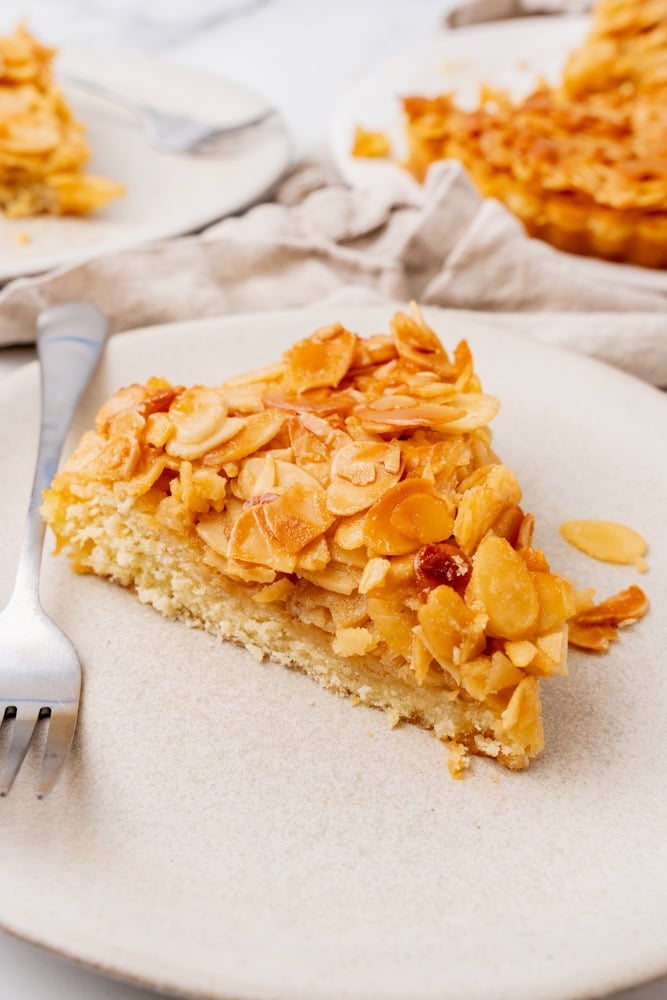 Side angle of a pie wedge shaped slice of almond tart on a beige plate with a fork.