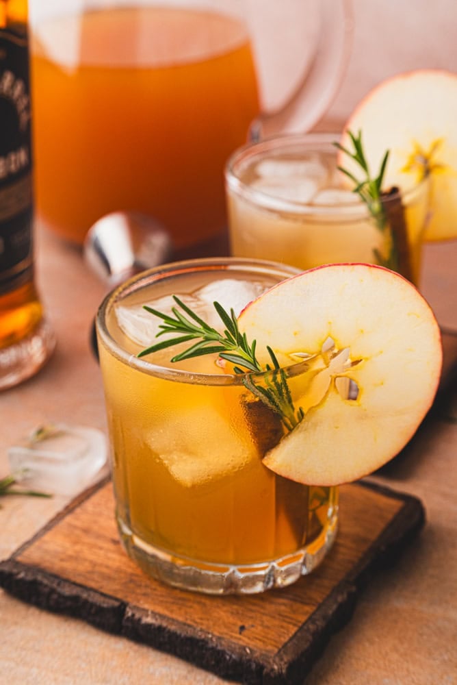 different angle of apple cider bourbon cocktail with apples and a little jar of maple syrup in the background. The top is garnished with a round apple slice, cinnamon stick, and rosemary. There's a pitcher of apple cider in the background.