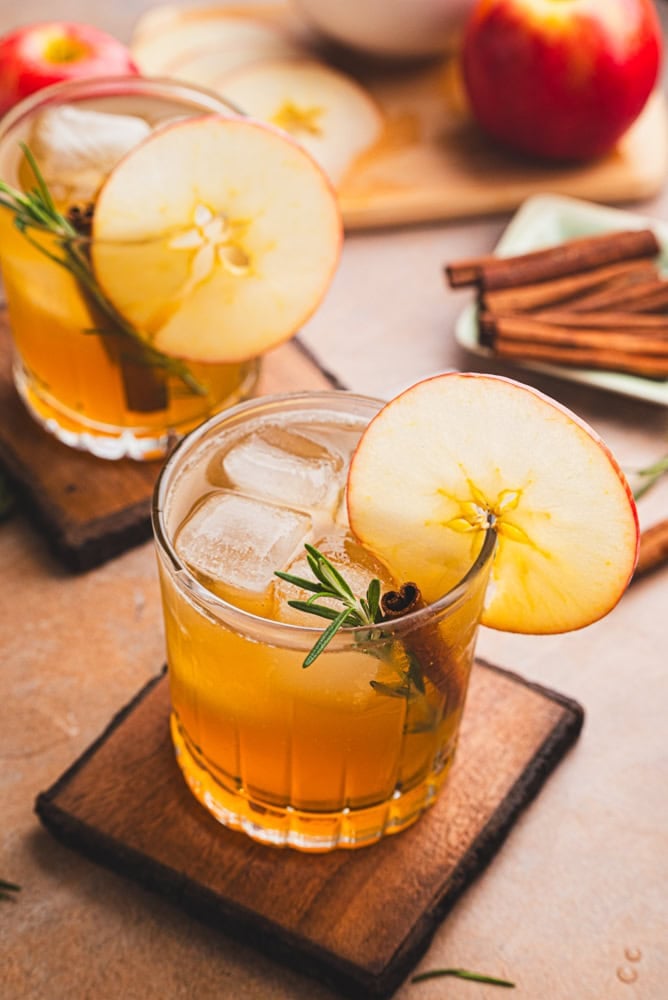 two apple cider bourbon cocktails with apples and a little jar of maple syrup in the background. The top is garnished with a round apple slice, cinnamon stick, and rosemary.